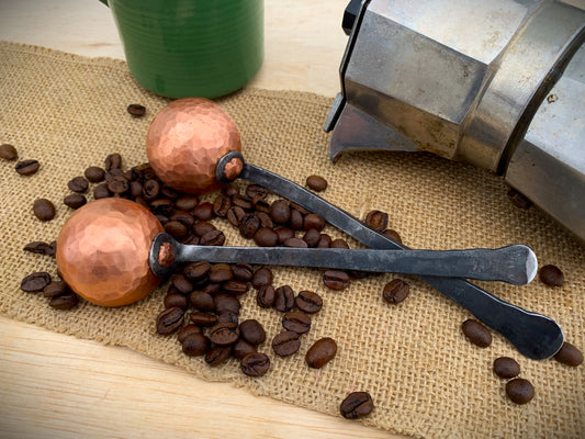 Copper coffee scoop on coffee beans with a green mug and metal container in the background.